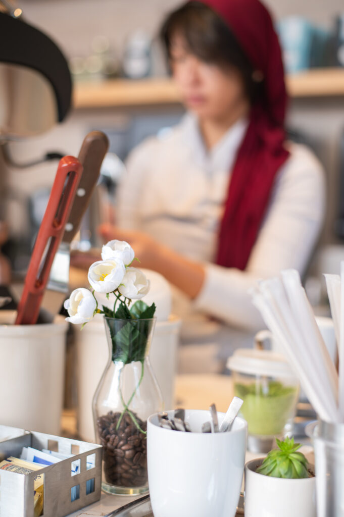 A vase of white faux flowers is in the foreground with a barista in a red scarf being in the background