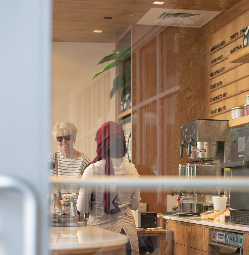 An elderly woman ordering coffee. She is smiling at the barista, wearing black shades, dangling earrings and a purse across her shoulder. 