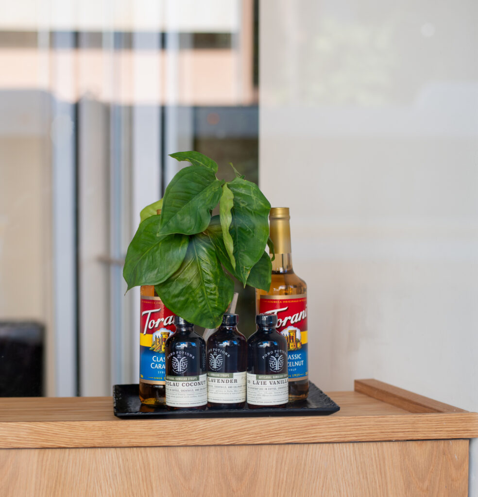 A window display of hawaiian made coffee syrups. A green plant is on the tray of syrup bottles behind them for decoration 