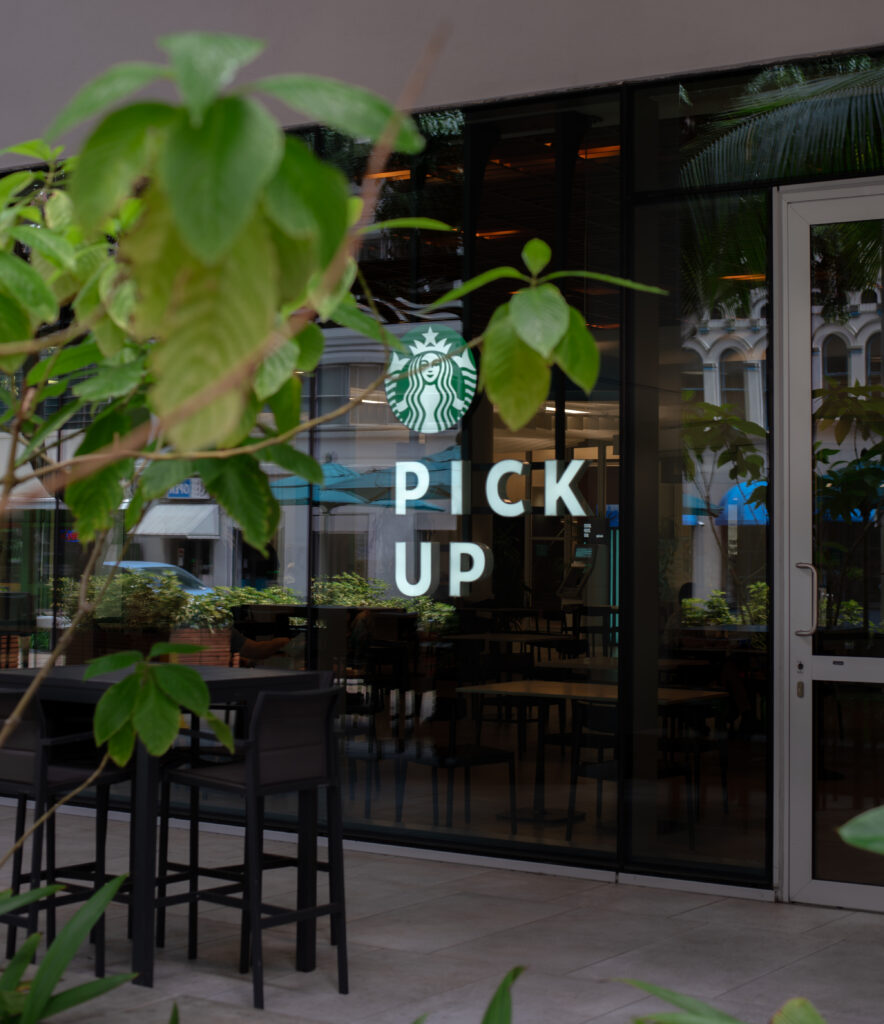 A store front advertising a Item pick up location, branches of a tree cover the store front a little, reflections of surrounding buildings can be seen in the glass