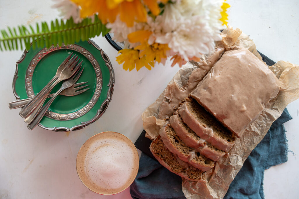 Pumpkin Spice Banana Bread on table with autumn flowers, a cafe latte and green pastry plates with silver forks. 